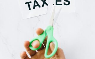 Top-down view of a hand holding scissors cutting the word 'taxes' on a marble surface. GST on Salon Serices has been cut down from 18% to 5%
